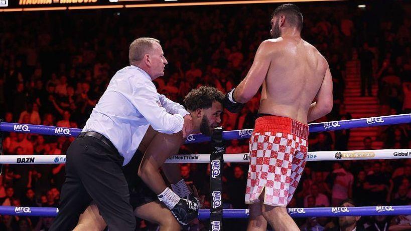 Moses Itauma being held on the ropes by the referee during a fight, as Filip Hrgovic observes the scene.