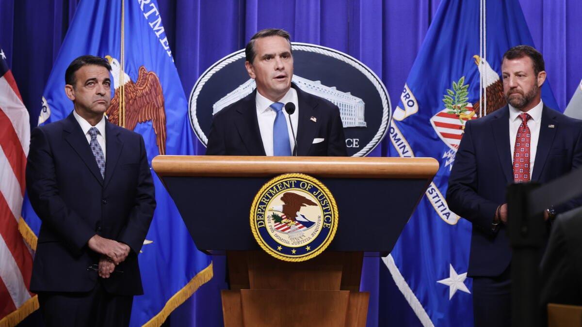 A man in a dark suit and blue tie addresses an audience from a lectern, with two other men in suits standing beside him.