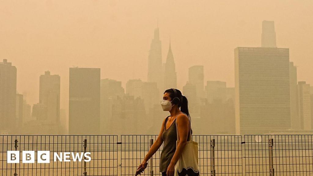 The New York City skyline, including prominent skyscrapers, is obscured by a thick orange haze from distant wildfires.